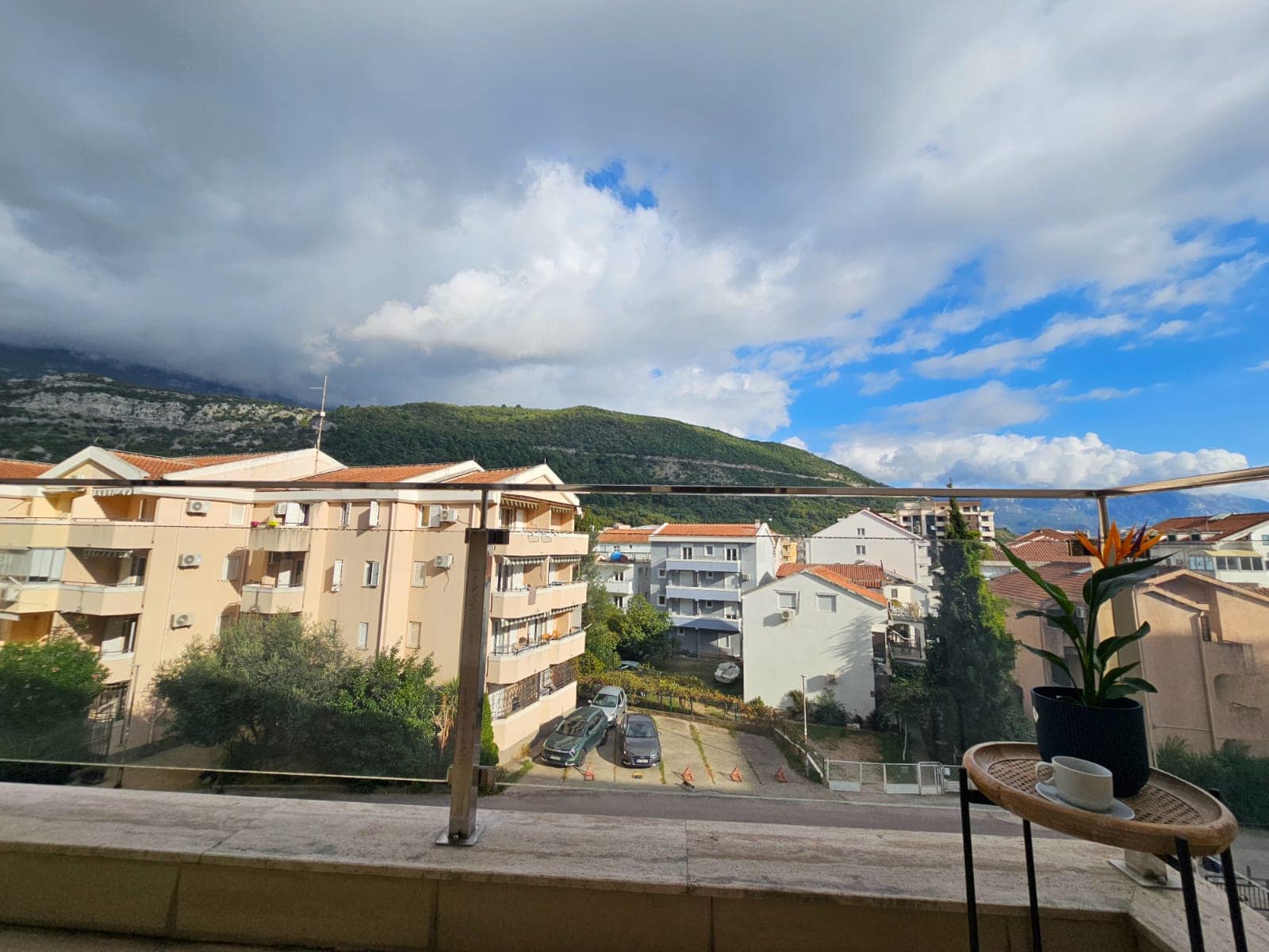 Pogled s balkona stana u Budvi, Crna Gora: panorama grada, planine i parking, stočić s cvećem u prvom planu.