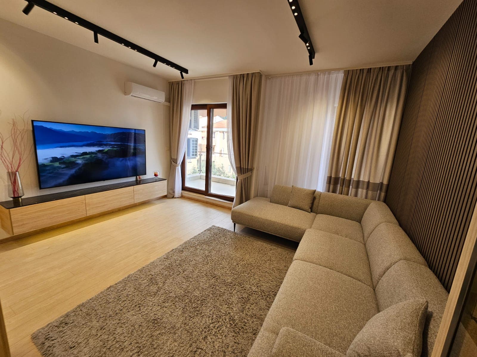 Savremena dnevna soba u stanu u Budvi, Crna Gora: ugaona garnitura, TV zona, rebrasti zid i izlaz na balkon.