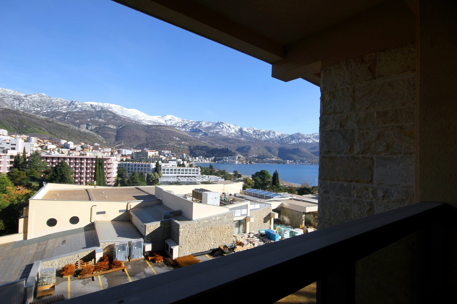 Blick vom Balkon der Wohnung in Budva, Montenegro: Panorama auf das Meer, die Stadt und schneebedeckte Berge an einem sonnigen Tag.