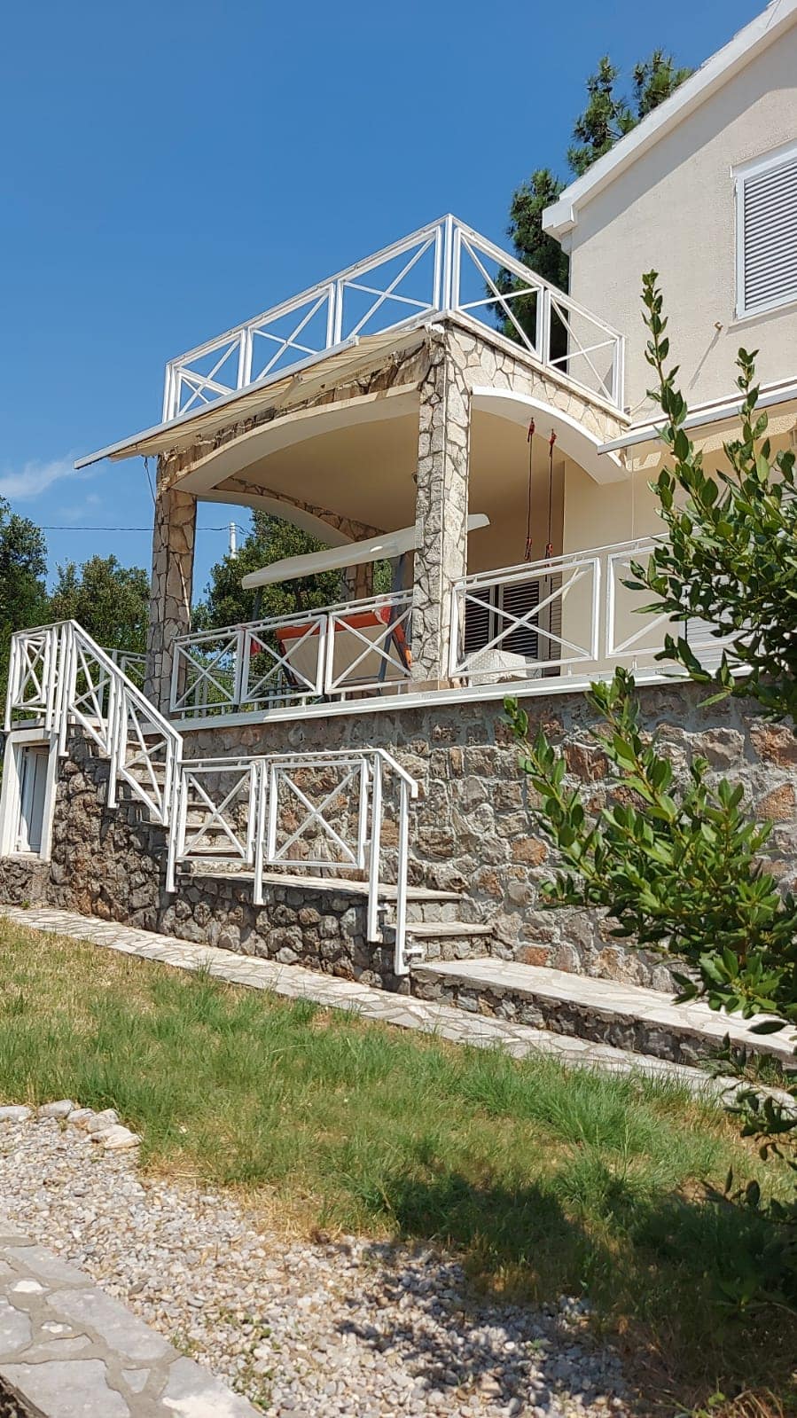 Villa in Bigovo, Montenegro: Hausfassade mit Steinverkleidung, großzügiger Terrasse und weißen Geländern vor dem Hintergrund eines Gartens.