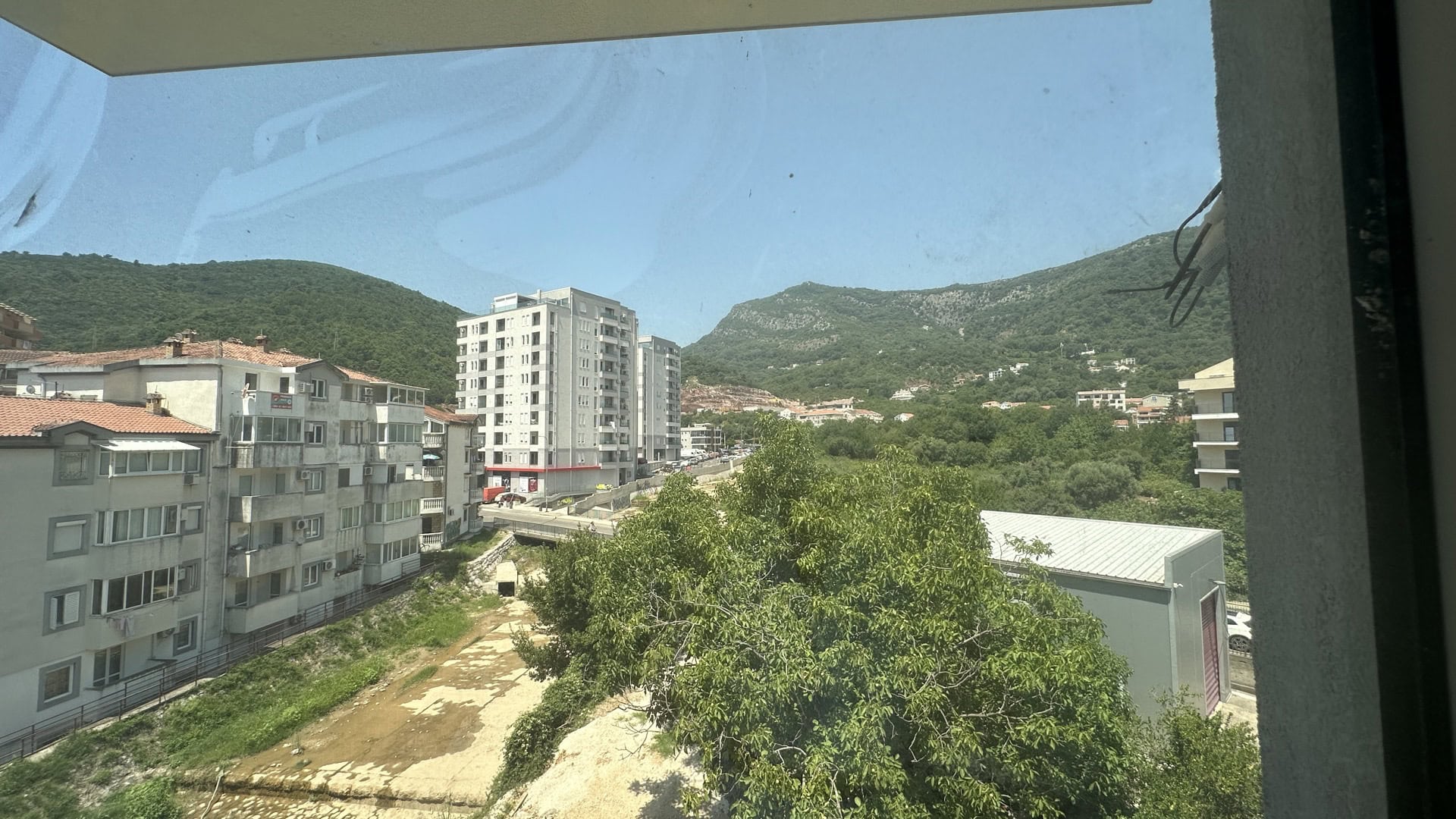Вид с балкона квартиры в Будве, Черногория: панорама на город, зеленые горы и жилые дома в солнечный день.