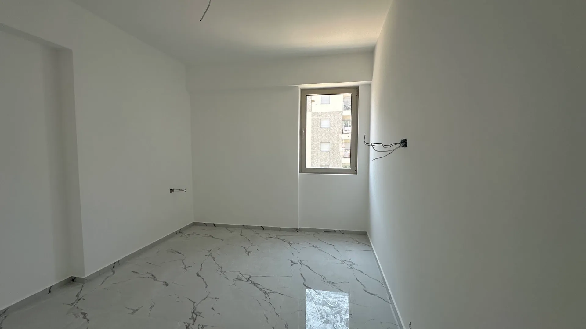 Helles Schlafzimmer in einem Neubau in Budva, Montenegro: weiße Wände, Feinsteinzeugboden in Marmoroptik, Blick aus dem Fenster.