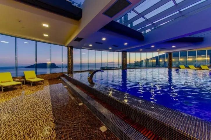 Überdachtes Schwimmbad mit Panoramablick auf das Meer in einer exklusiven Wohnanlage in Budva, Montenegro, mit Ruhebereich und Liegestühlen.