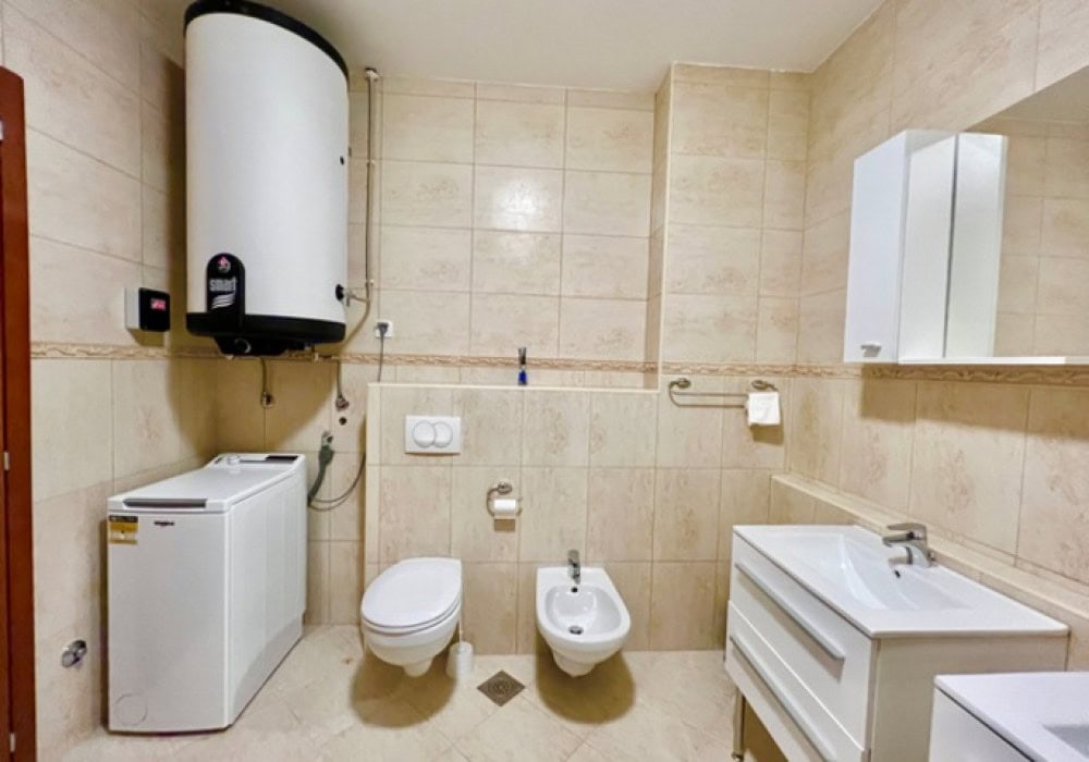 Badezimmer in einer Wohnung in Bečići, Montenegro: beige Fliesen, Boiler, Waschmaschine, Sanitäranlagen und Waschtisch mit Waschbecken.