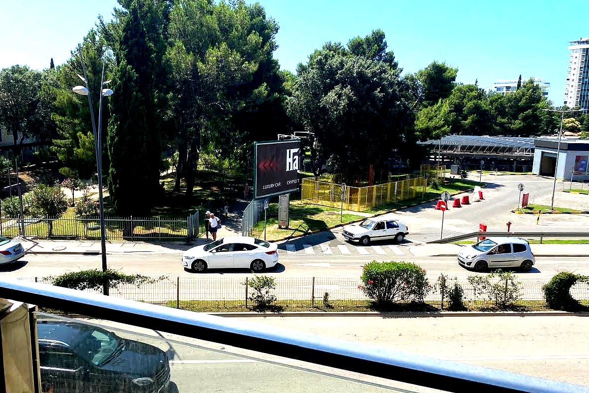 Blick vom Balkon einer Wohnung im Zentrum von Budva, Montenegro: Stadtstraße, grüner Park und Parkplatz neben dem Haus.
