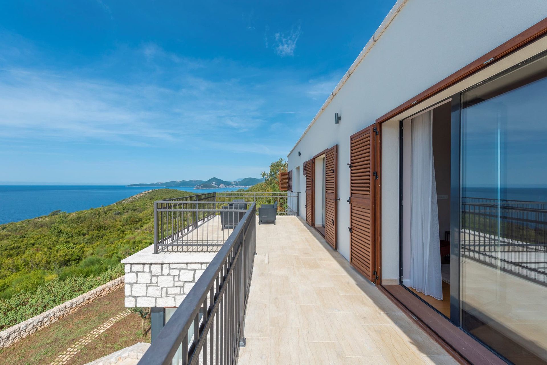 Spacieuse terrasse d’une maison à Reževići, Monténégro, avec vue panoramique sur la mer, parement en pierre et volets en bois.