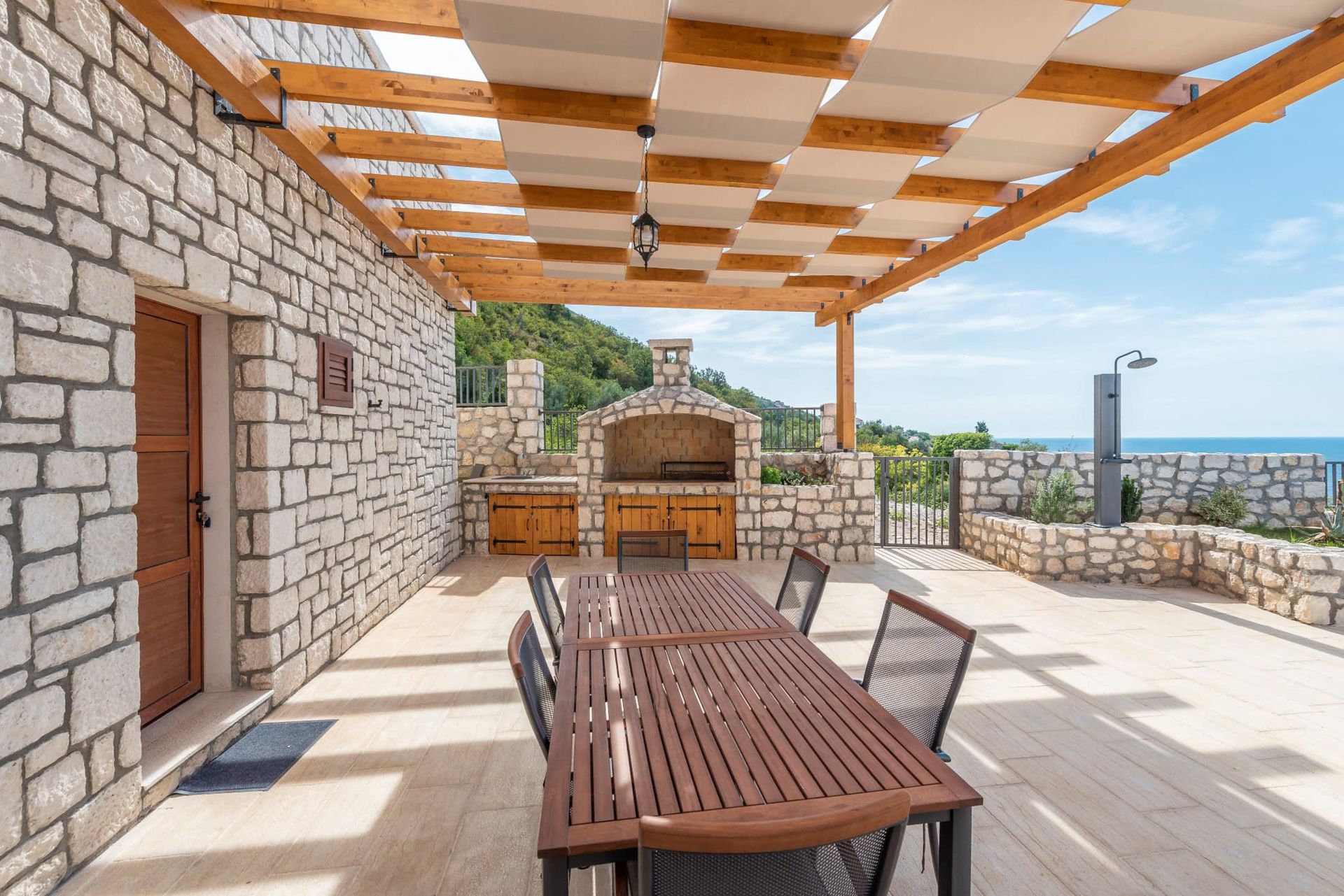 Terrasse de la maison à Reževići, Monténégro : revêtement en pierre, barbecue, table en bois et vue sur la mer sous un auvent.