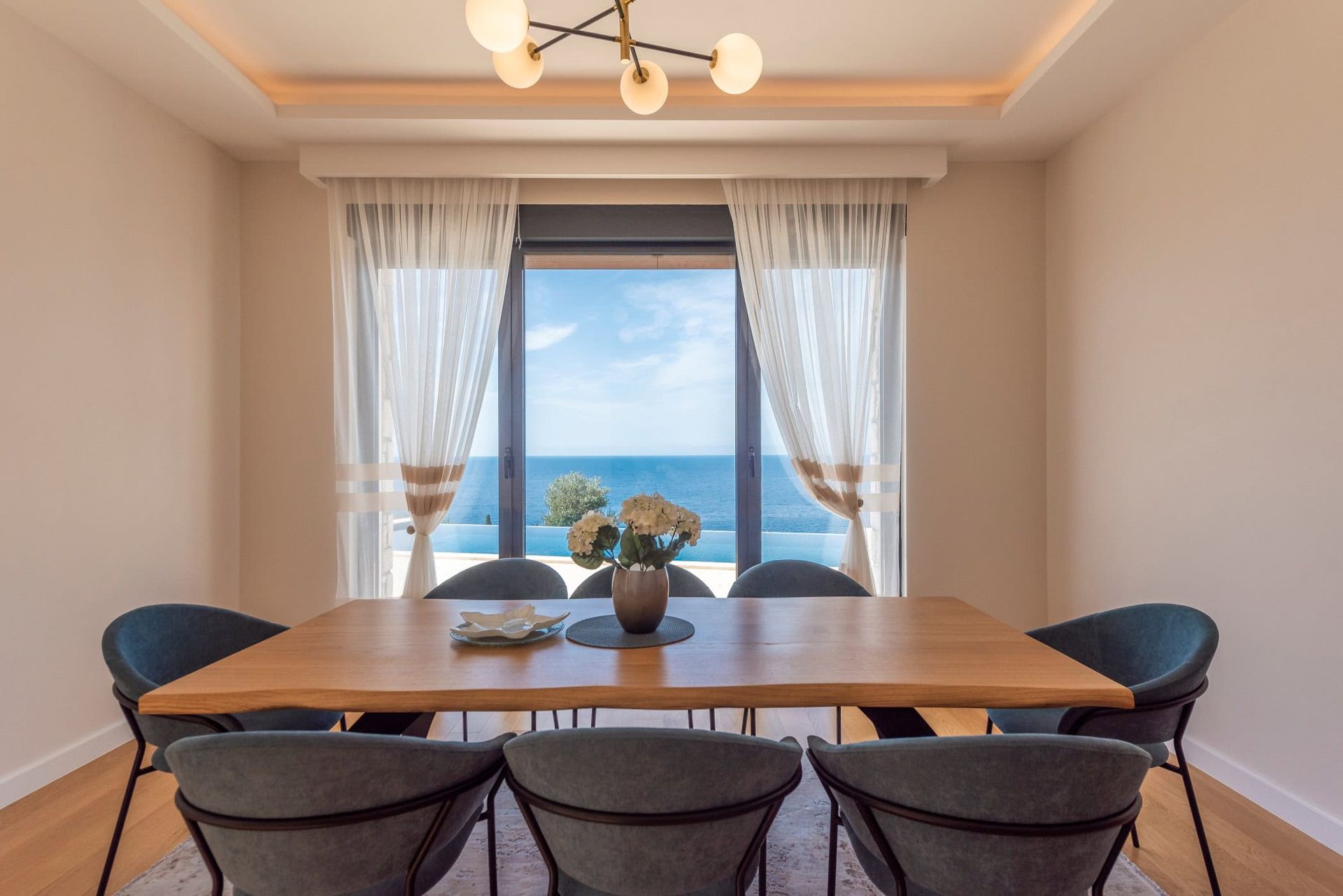 Coin repas dans une maison à Reževići, Monténégro : table en bois, chaises grises et vue panoramique sur la mer depuis la fenêtre.