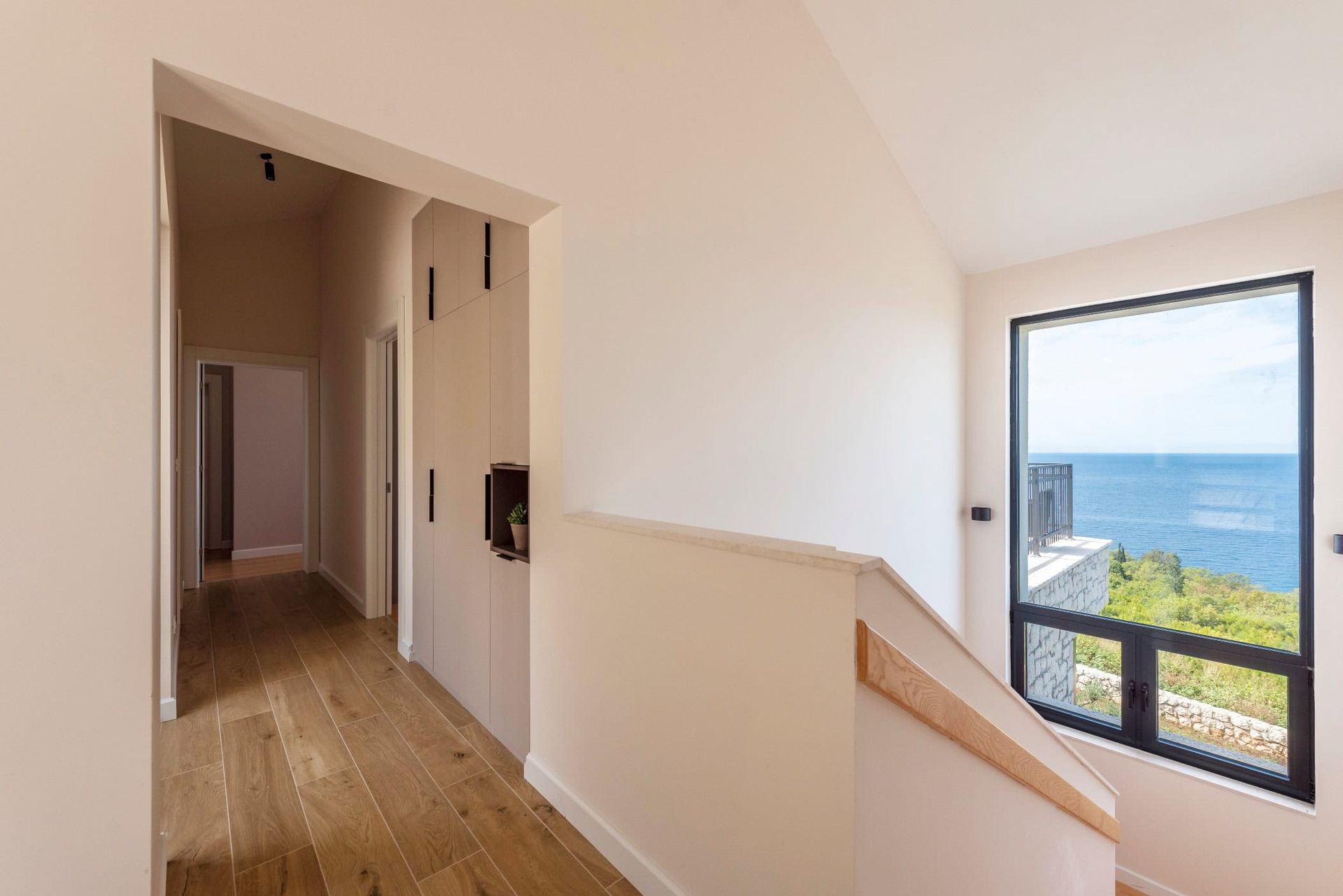 Intérieur d’une maison à Reževići, Monténégro : couloir lumineux avec baie vitrée panoramique et vue sur la mer, placard intégré et escalier.