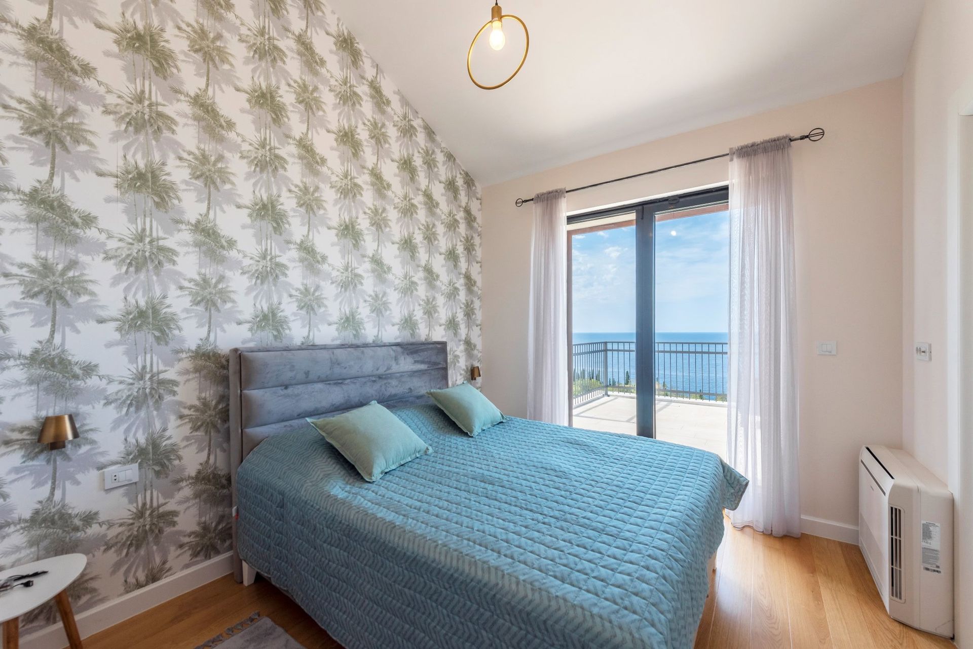 Chambre dans une maison à Reževići, Monténégro : lit double, papier peint avec des palmiers et vue panoramique sur la mer depuis la terrasse.
