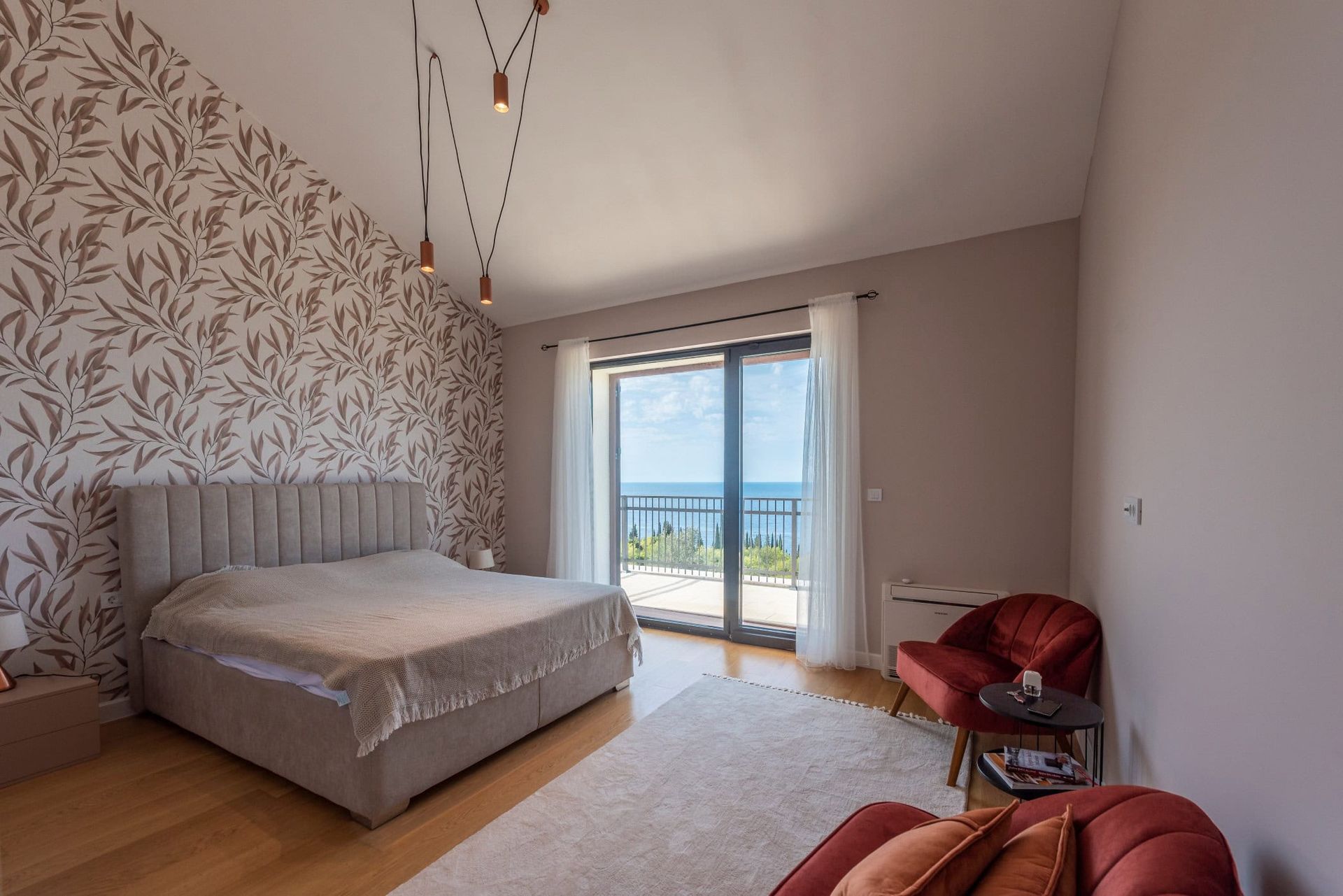 Chambre dans une maison à Reževići, Monténégro : lit double, mur d’accent, fauteuils rouges et vue sur la mer depuis la terrasse.