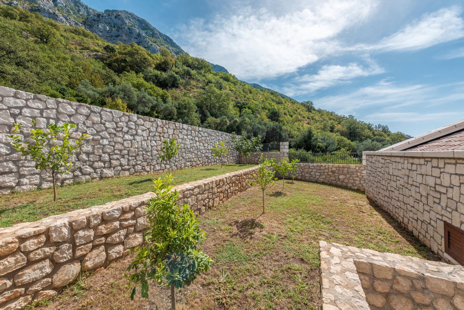 Jardin bien entretenu avec terrasses en pierre et jeunes arbres près de la maison à Reževići, Monténégro, vue sur les montagnes.