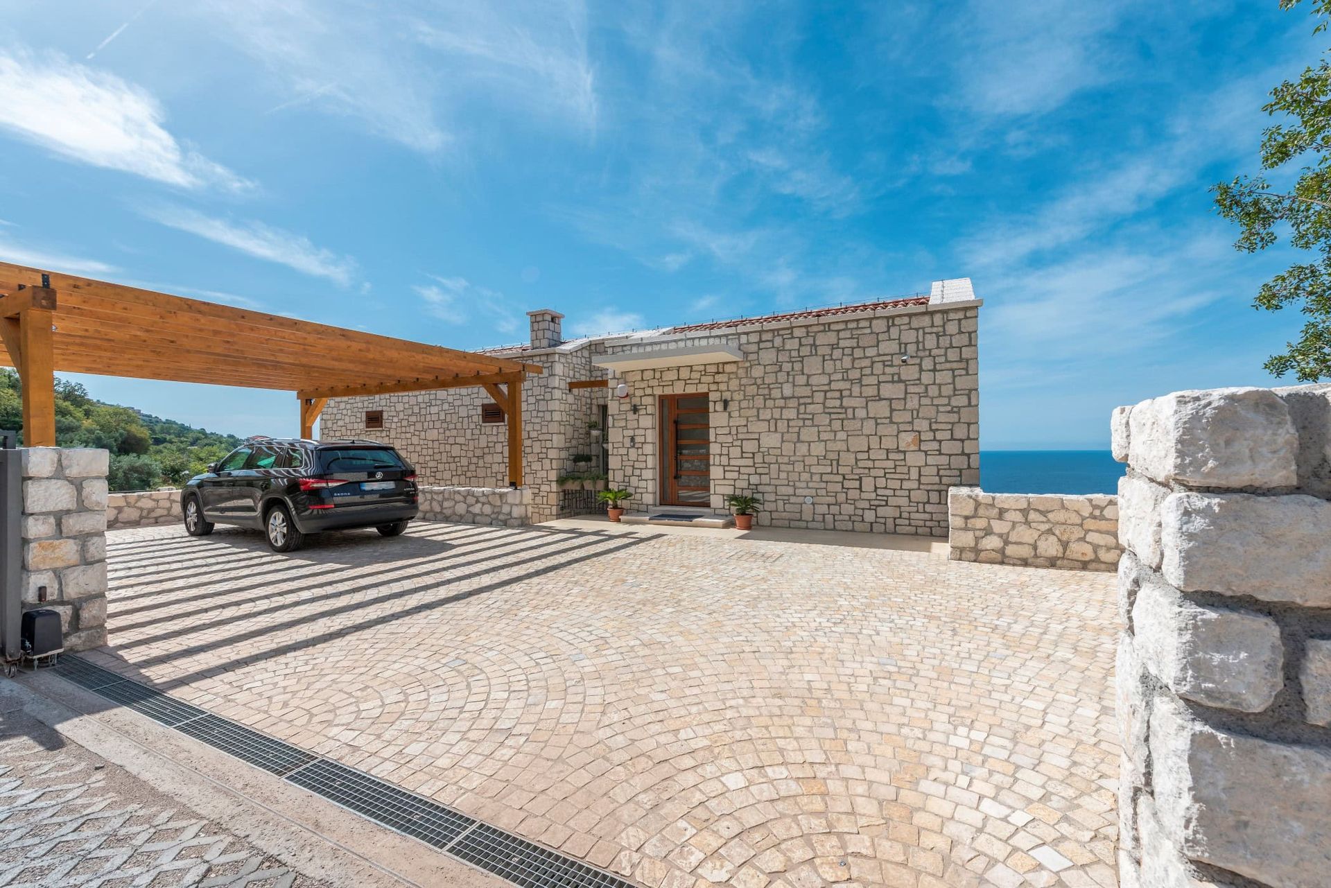 Maison en pierre à Reževići, Monténégro : façade en pierre naturelle, parking avec abri en bois et vue sur la mer.