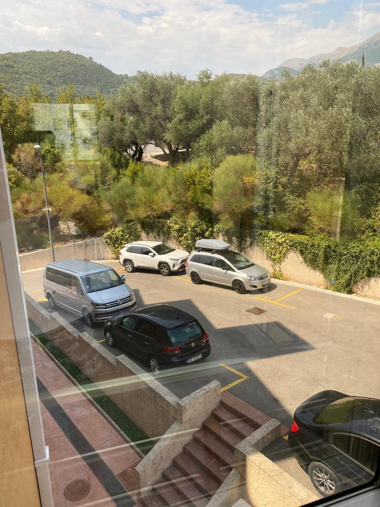 Blick aus dem Fenster einer Wohnung in Bečići, Montenegro: Parkplatz am Haus, grüne Bäume und Berglandschaft im Hintergrund.