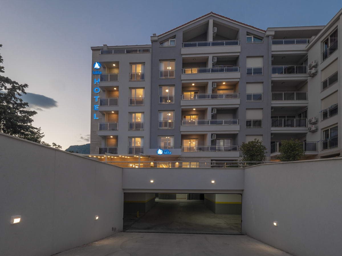 Fassade des Hotels Idila in Budva, Montenegro, mit modernen Balkonen und Zufahrt zur Tiefgarage bei Abenddämmerung.