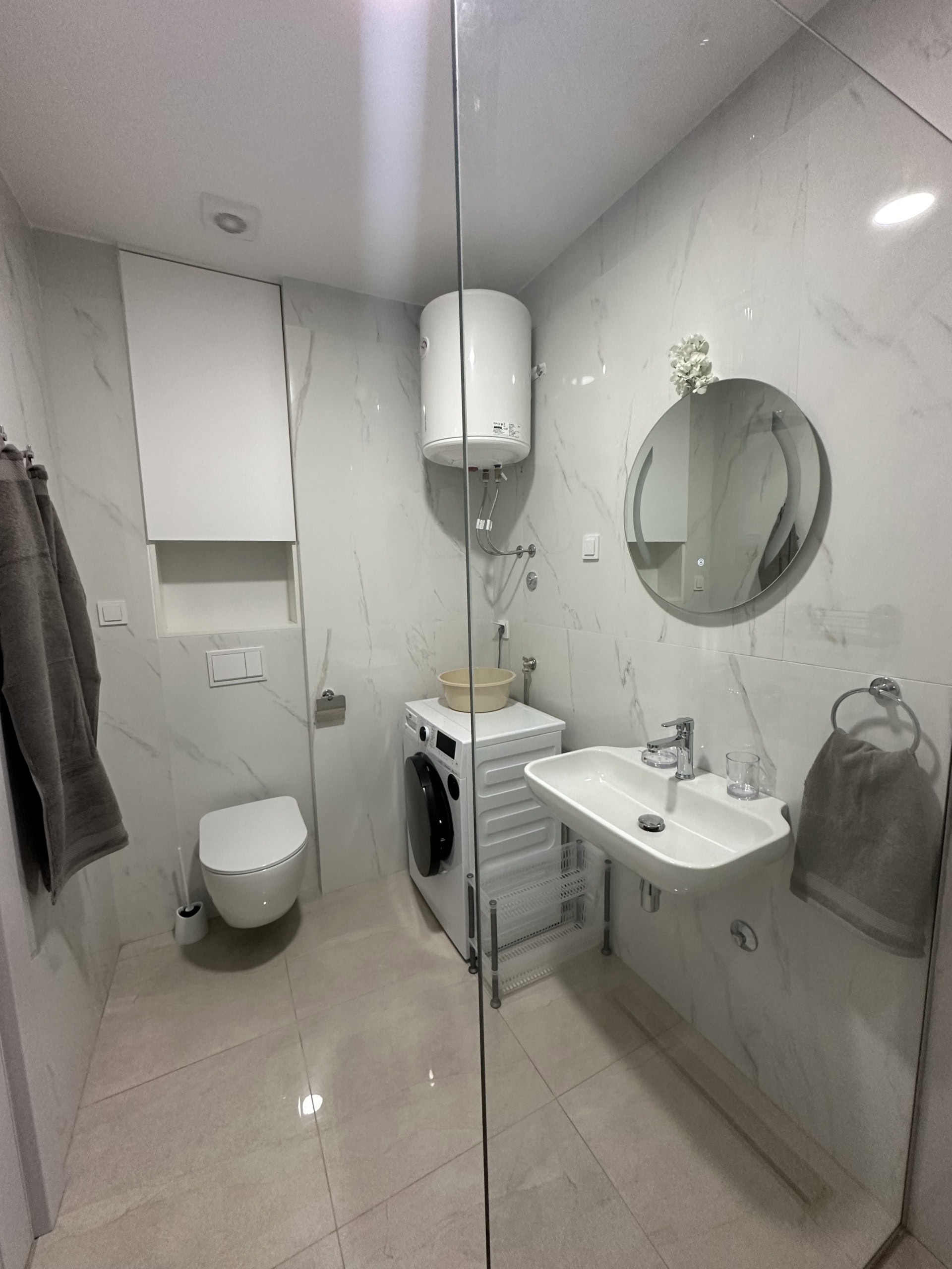 Modernes Badezimmer in einer Wohnung in Bečići, Montenegro: helle Fliesen, wandhängendes WC, Waschbecken und Waschmaschine.