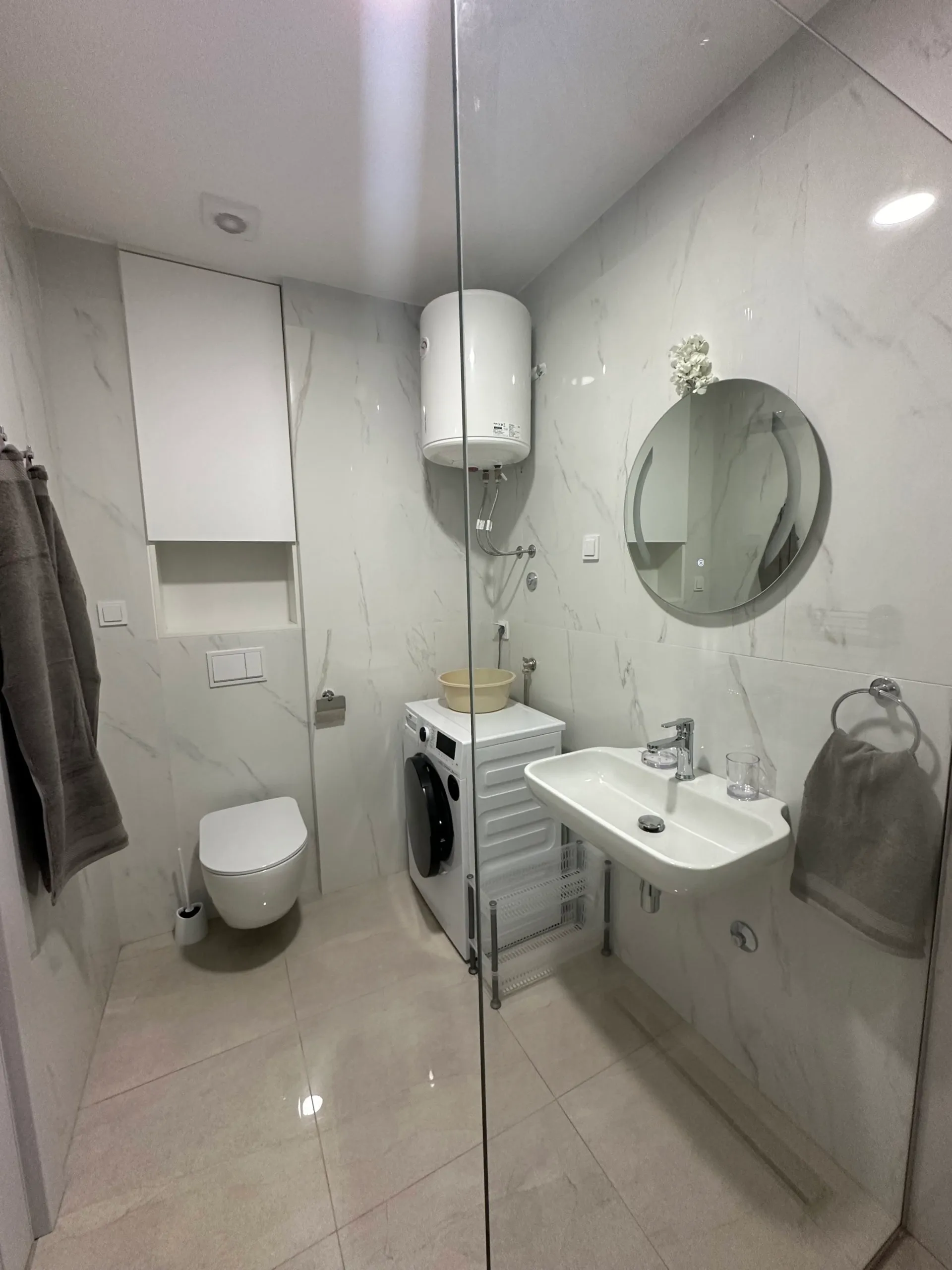 Modernes Badezimmer in einer Wohnung in Bečići, Montenegro: helle Fliesen, wandhängendes WC, Waschbecken und Waschmaschine.