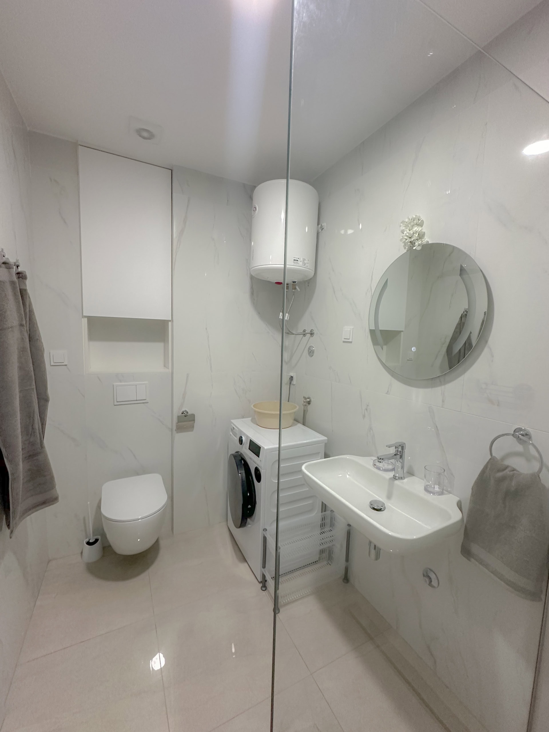 Modernes Badezimmer in einer Wohnung in Bečići, Montenegro: helle Fliesen, Hänge-WC, Waschbecken und Waschmaschine.