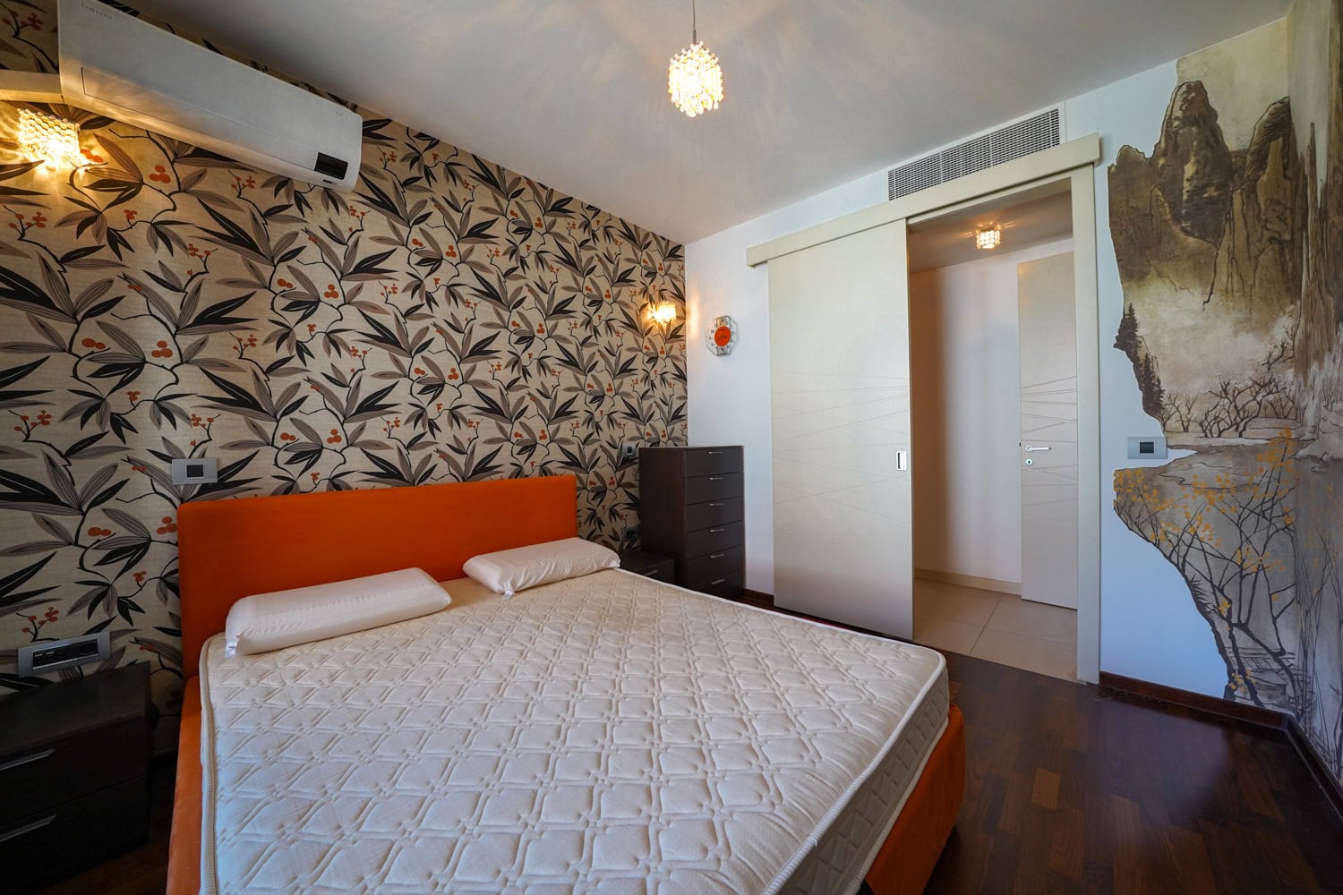 Schlafzimmer in einer Wohnung in Budva, Montenegro: orangefarbenes Bett, Tapete mit Pflanzenmuster und Schiebetüren.