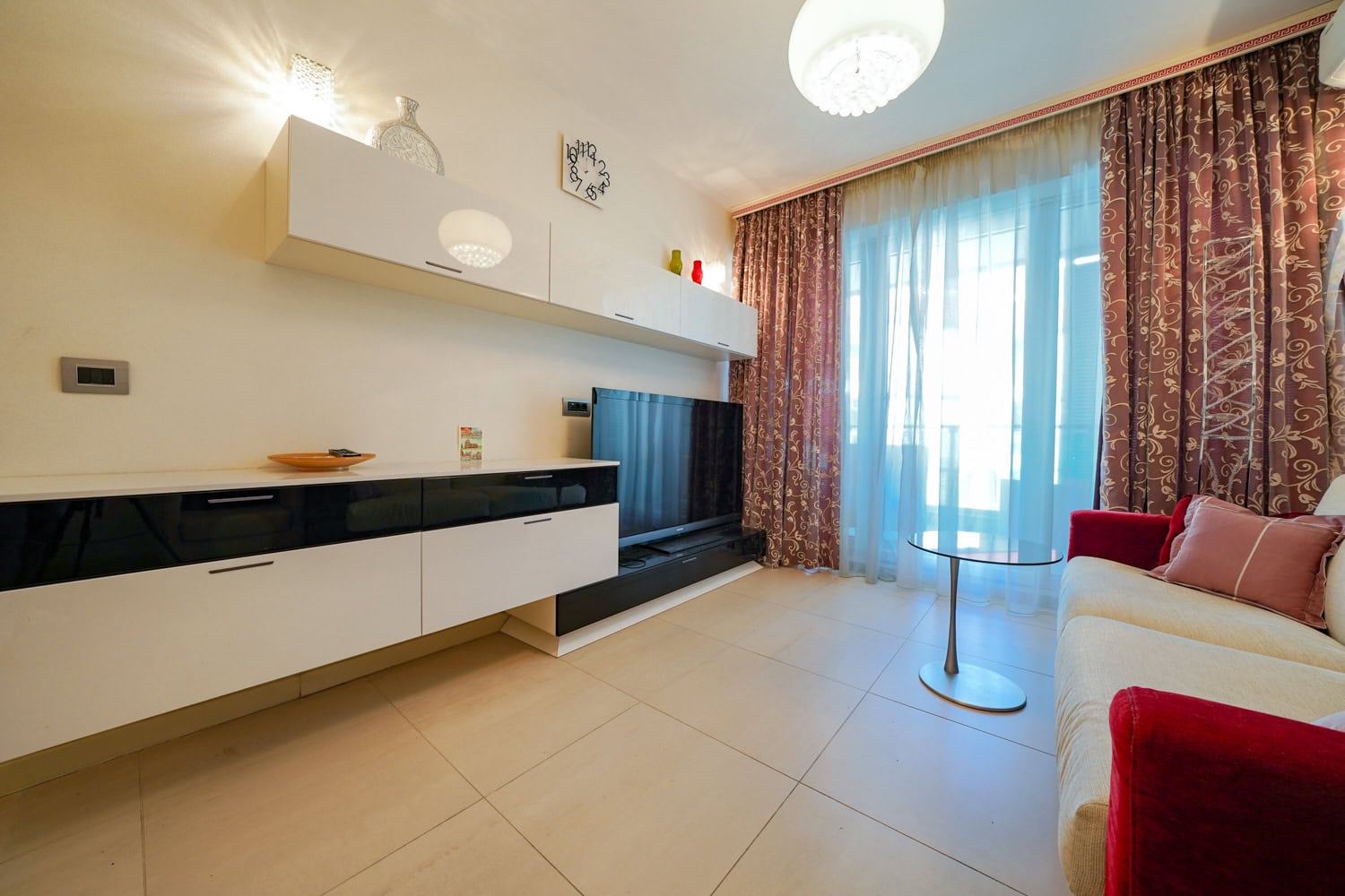 Wohnzimmer in einer Wohnung in Budva, Montenegro: moderne Möbel, TV-Bereich, helle Fliesen und ein Panoramafenster mit Vorhängen.