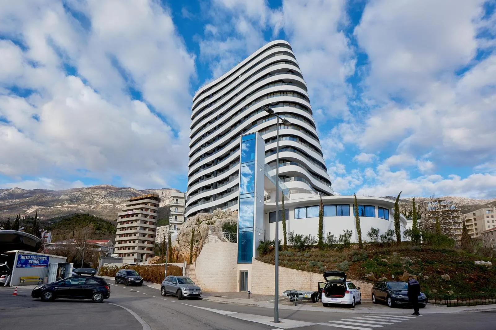 Exterior of a modern white high-rise building with wavy balconies, set against mountains and sky.