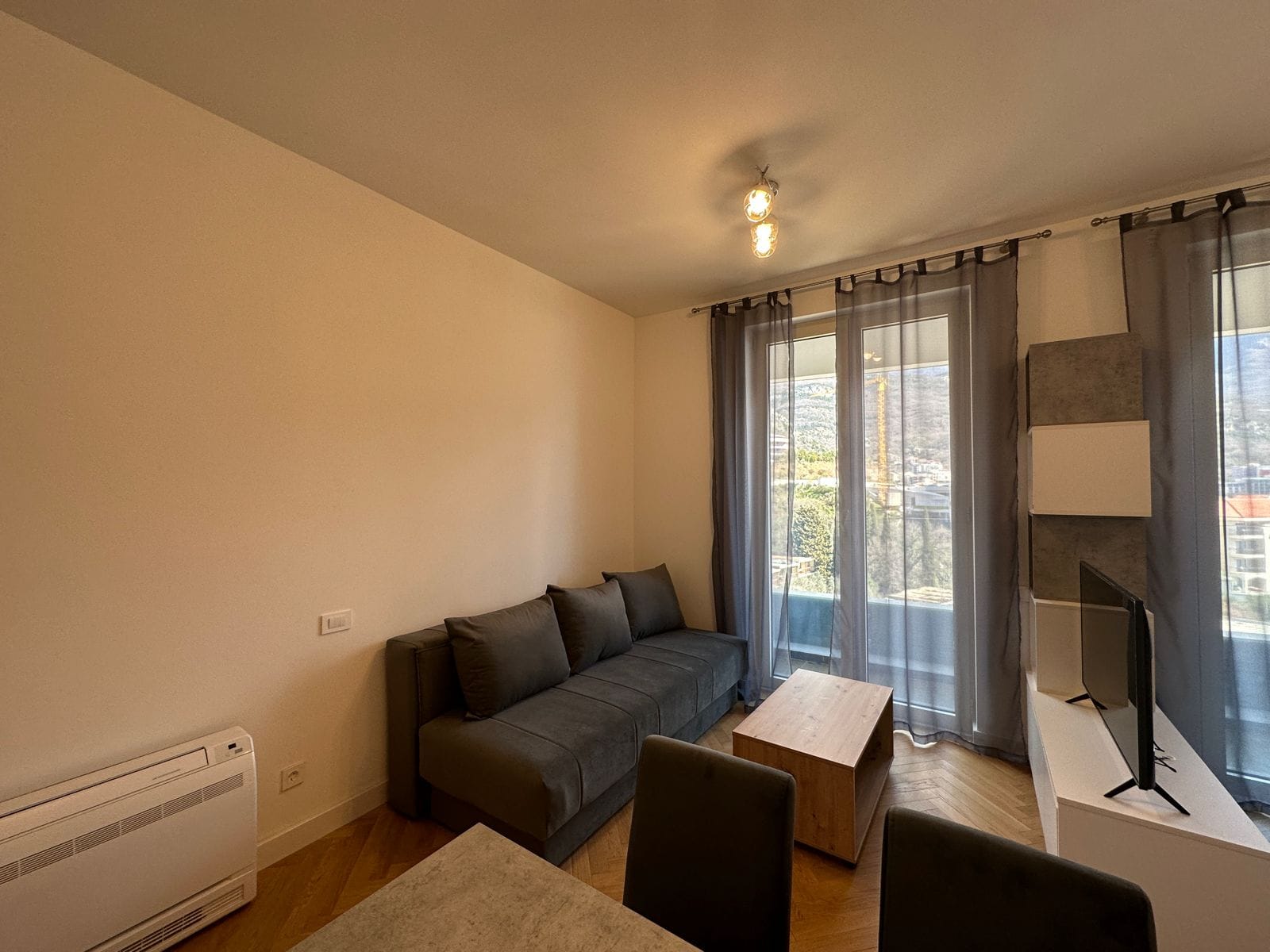 Salon w nowoczesnym mieszkaniu w Bečići, Czarnogóra: szara sofa, drewniany stolik i wyjście na balkon z widokiem.