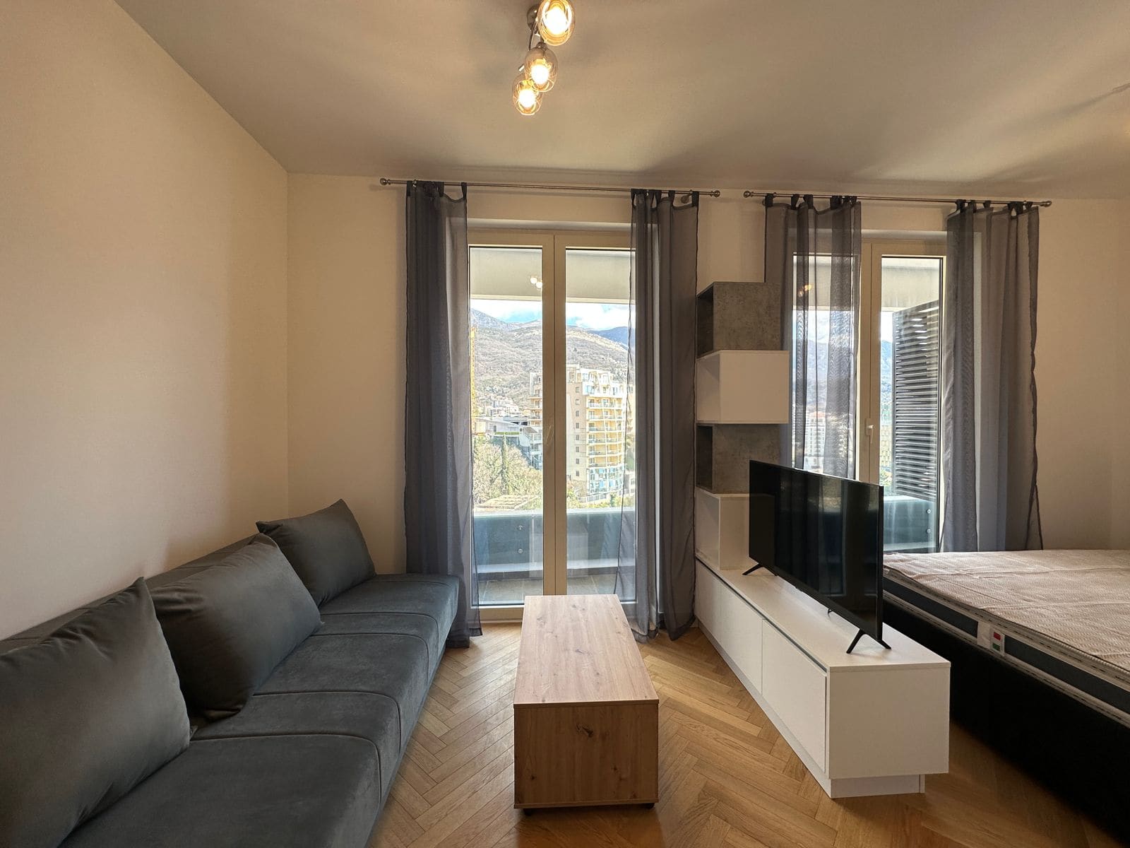 Salon w mieszkaniu w Bečići, Czarnogóra: szara sofa, strefa TV, panoramiczne okna z widokiem na góry i parkiet.