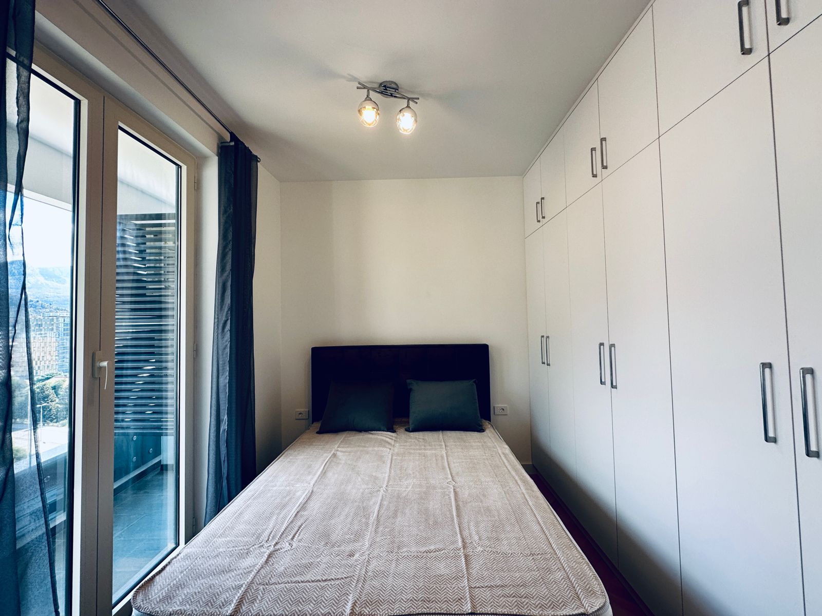 Sypialnia z łóżkiem dwuosobowym i wbudowaną szafą w mieszkaniu w Bečići, Czarnogóra, z wyjściem na balkon.