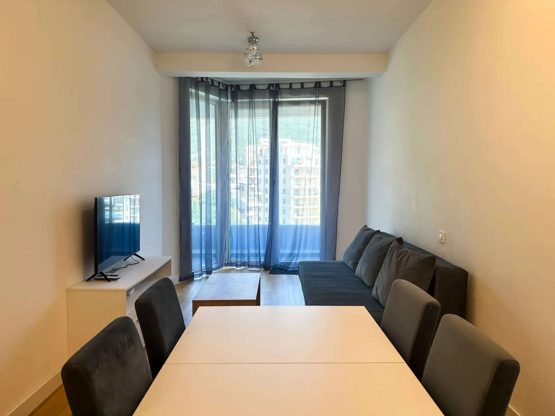 Wohnzimmer mit grauem Sofa, weißem Esstisch, Stühlen und Fernseher vor einem Fenster mit blauen Vorhängen.