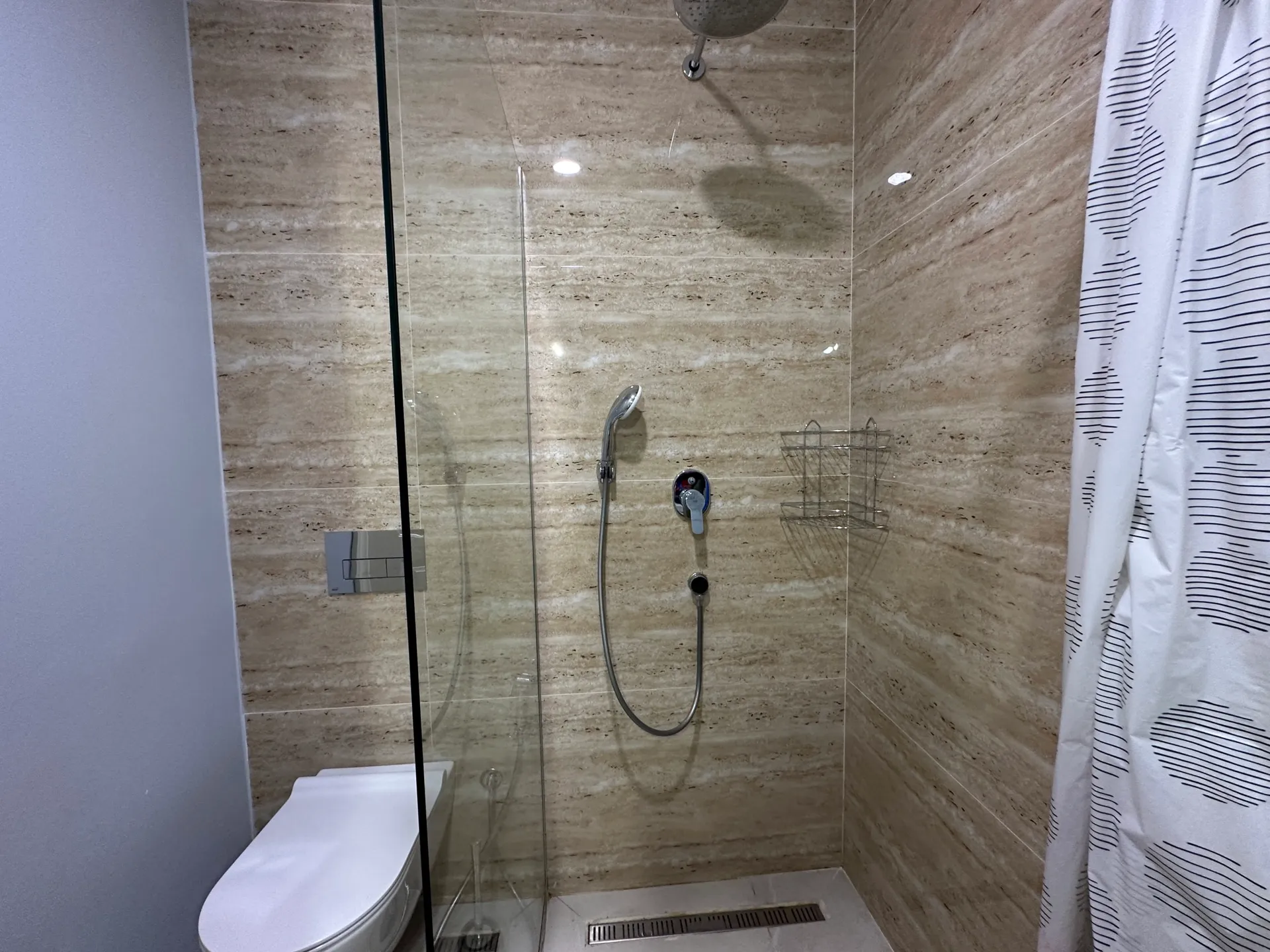 Badezimmer mit Marmor-Dusche, Glasabtrennung, wandmontierter Toilette und weißem Muster-Vorhang.