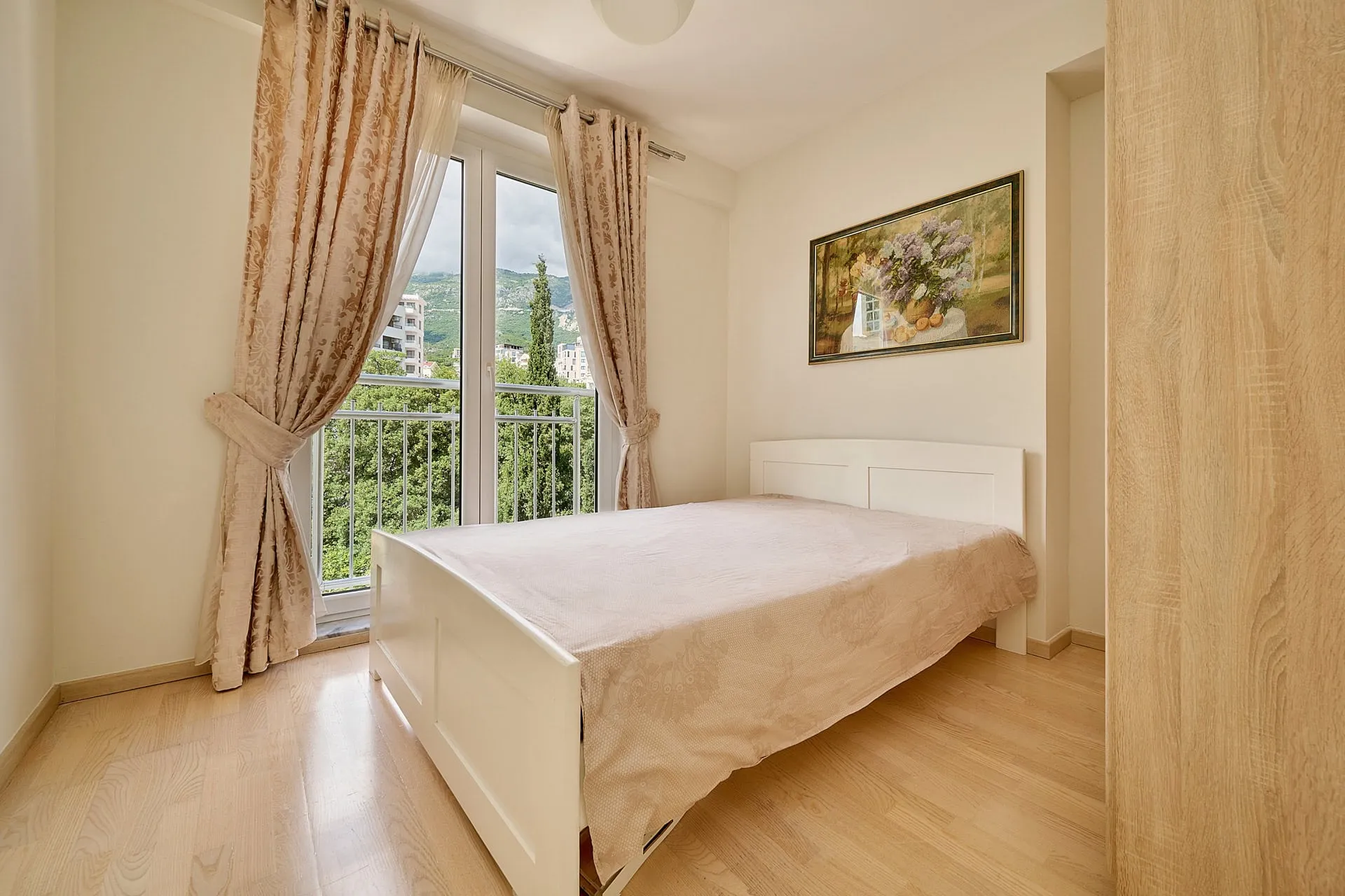 Schlafzimmer mit weißem Bett und Bergblick in einer Wohnung in Bečići, Montenegro: helles Interieur, Parkett, Panoramafenster.