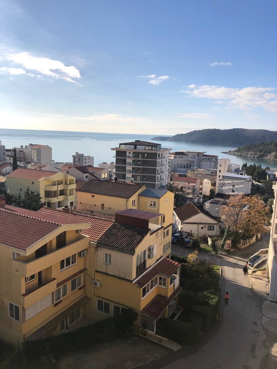 Panoramski pogled na more i grad Bečići, Crna Gora, iz prozora stana sa krovovima od crepa i planinskim pejzažem.