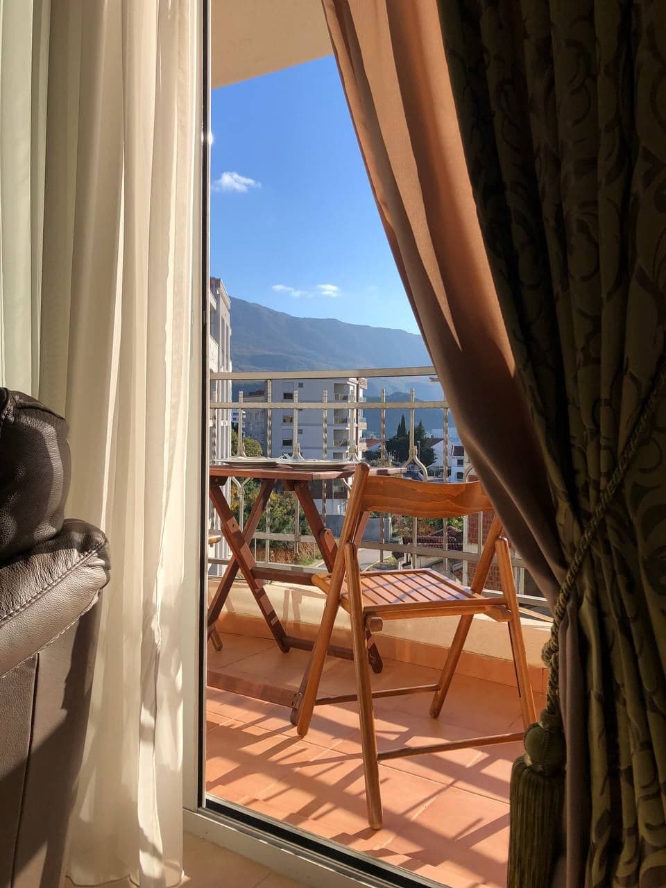 Pogled sa balkona stana u Bečićima, Crna Gora: drveni nameštaj, panoramski pogled na planine i grad kroz otvorena vrata.