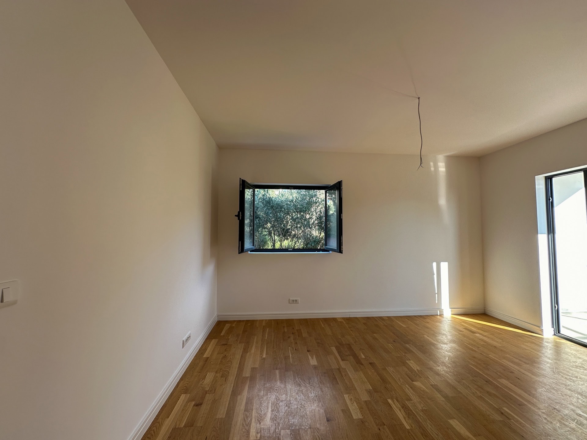 Helles Wohnzimmer mit Parkett in einer neuen Wohnung in Bečići, Montenegro, Blick ins Grüne aus dem offenen Fenster.