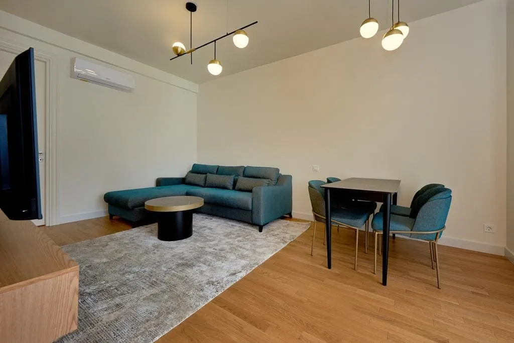 Modern living room featuring a blue sectional sofa, dining table set, TV, and pendant lights on light walls with wood flooring.