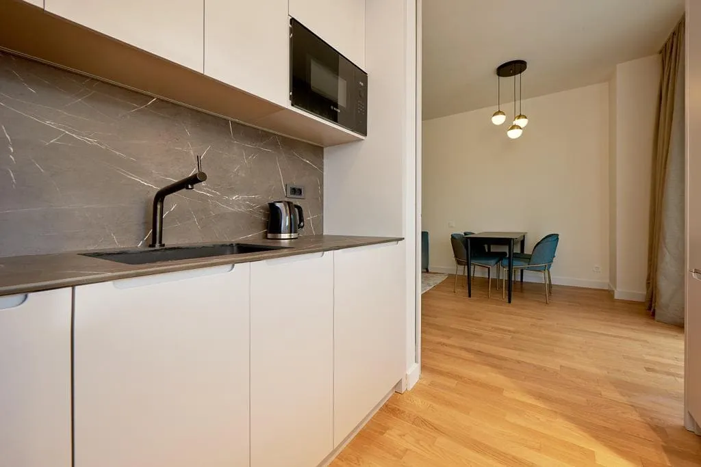 Modern kitchen with white cabinets, grey countertop, microwave, sink, and dining area visible.