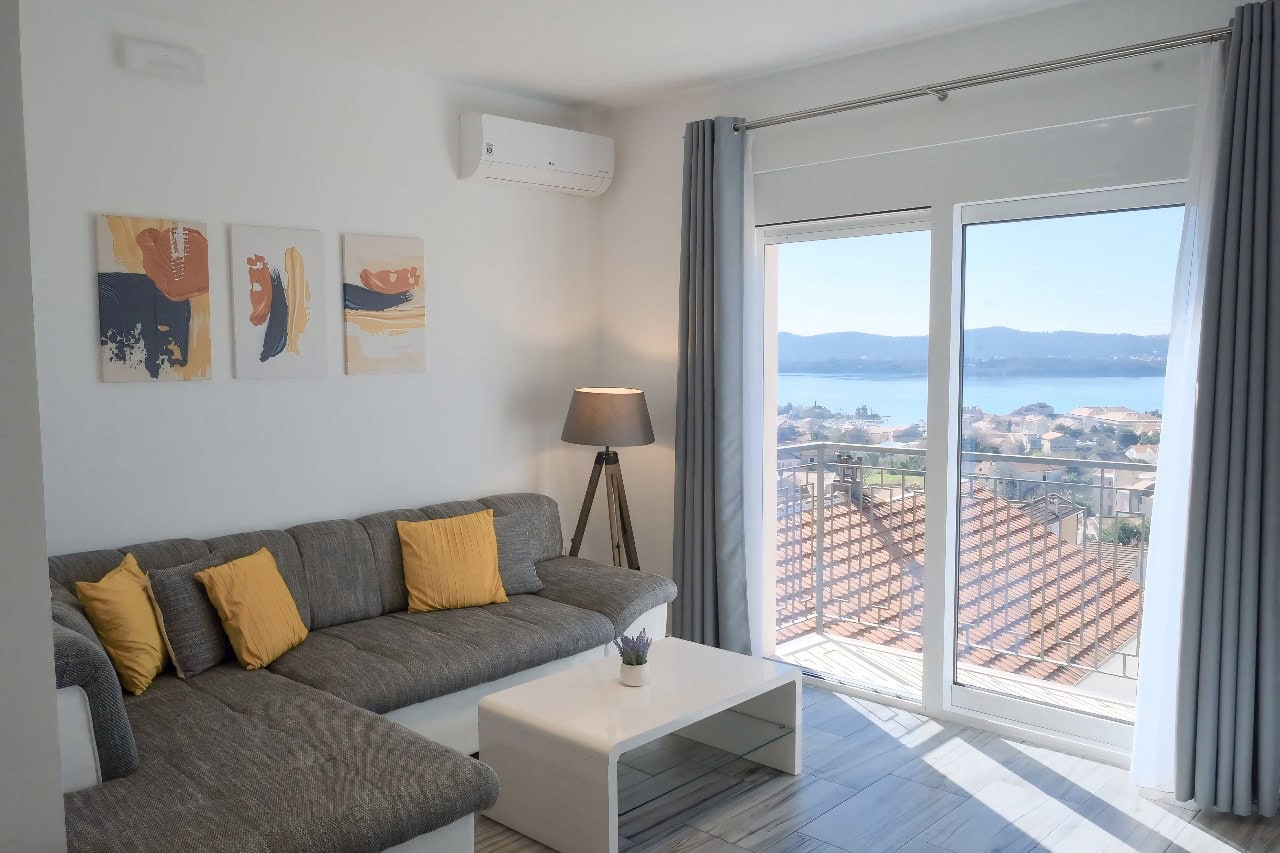 Helles Wohnzimmer in einer Wohnung in Tivat, Montenegro: graues Sofa, Panoramablick auf das Meer und Balkon mit Terrakottadach.