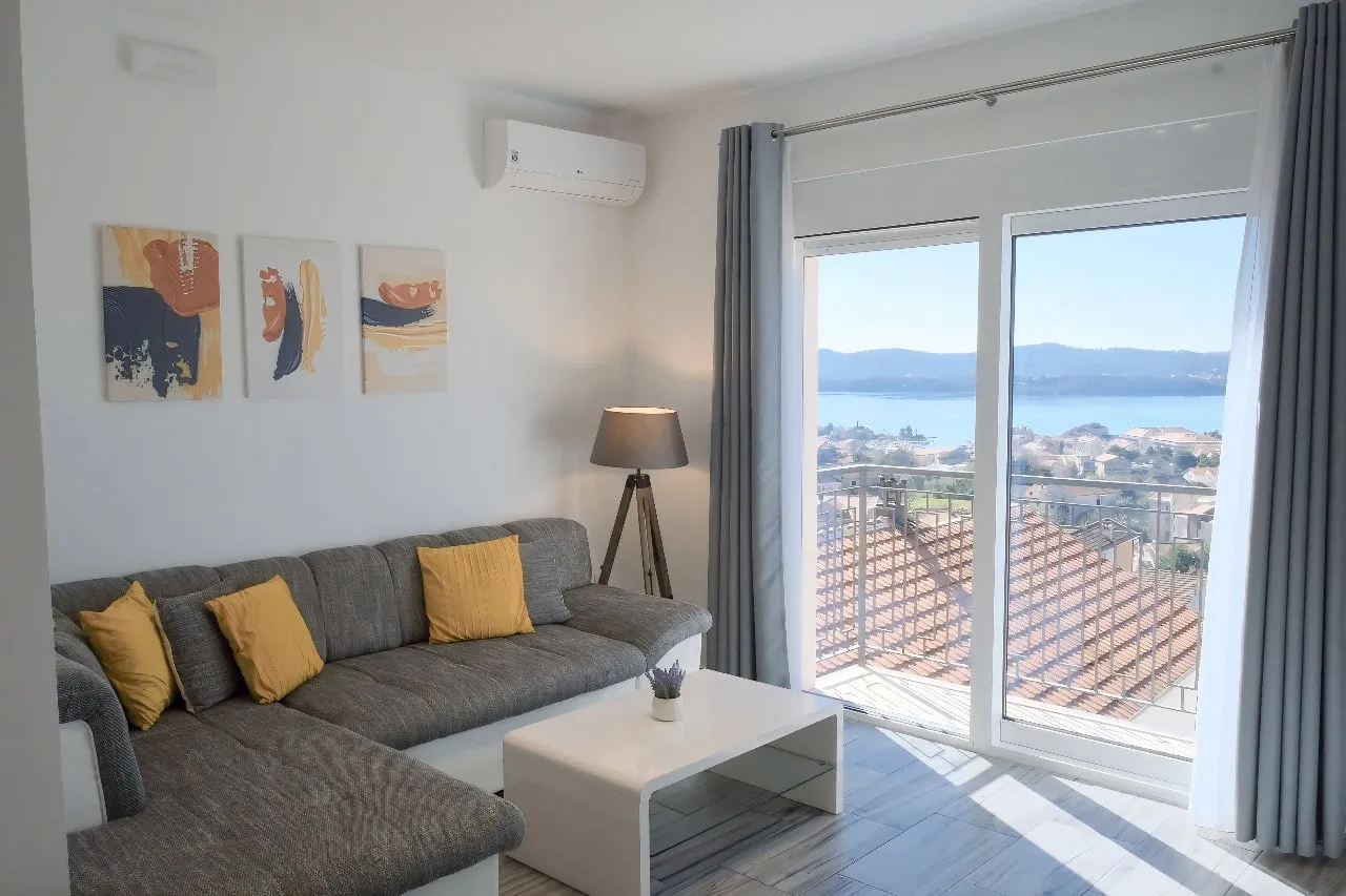 Helles Wohnzimmer in einer Wohnung in Tivat, Montenegro: graues Sofa, Panoramablick auf das Meer und Balkon mit Terrakottadach.