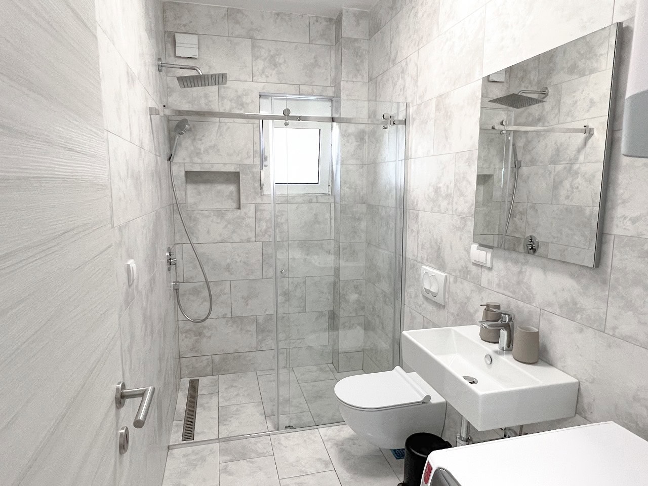 Modernes Badezimmer mit Duschkabine in einer Wohnung, Tivat, Montenegro: helle Fliesen, wandhängendes WC und Waschbecken.
