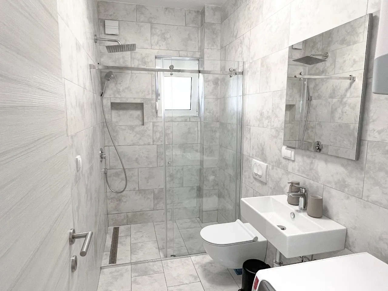 Modernes Badezimmer mit Duschkabine in einer Wohnung, Tivat, Montenegro: helle Fliesen, wandhängendes WC und Waschbecken.
