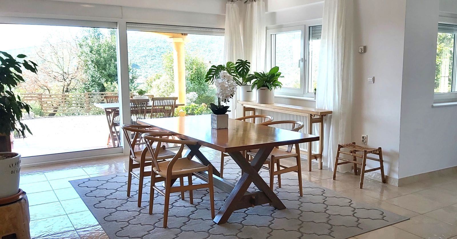 Kırımovitsa, Karadağ’daki evde yemek alanlı aydınlık oturma odası: ahşap masa, panoramik pencereler ve terasa manzara.