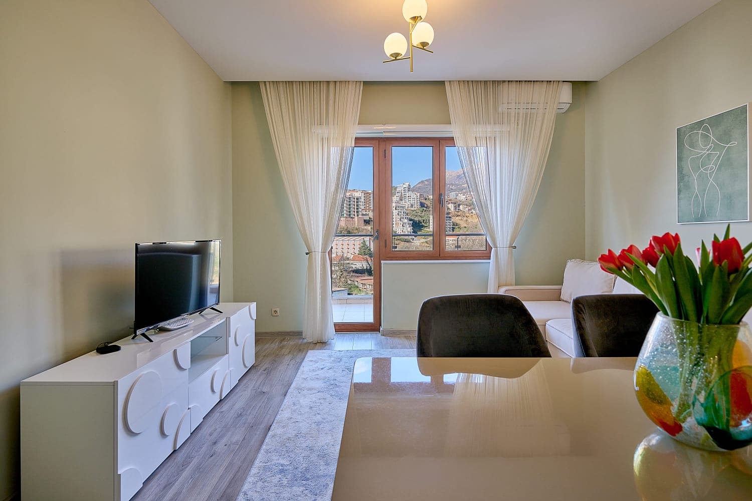 Ugodan dnevni boravak u stanu u Bečićima, Crna Gora: svetao nameštaj, TV zona i izlaz na balkon sa pogledom na planine.