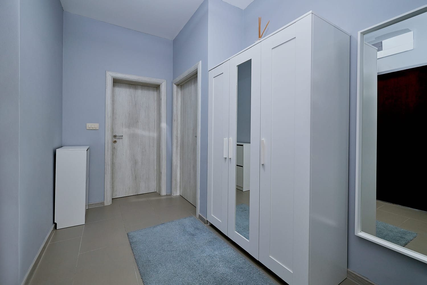 Svetlo predsoblje u stanu u Bečićima, Crna Gora: beli ormar sa ogledalom, siva vrata i pod od pločica u enterijeru.