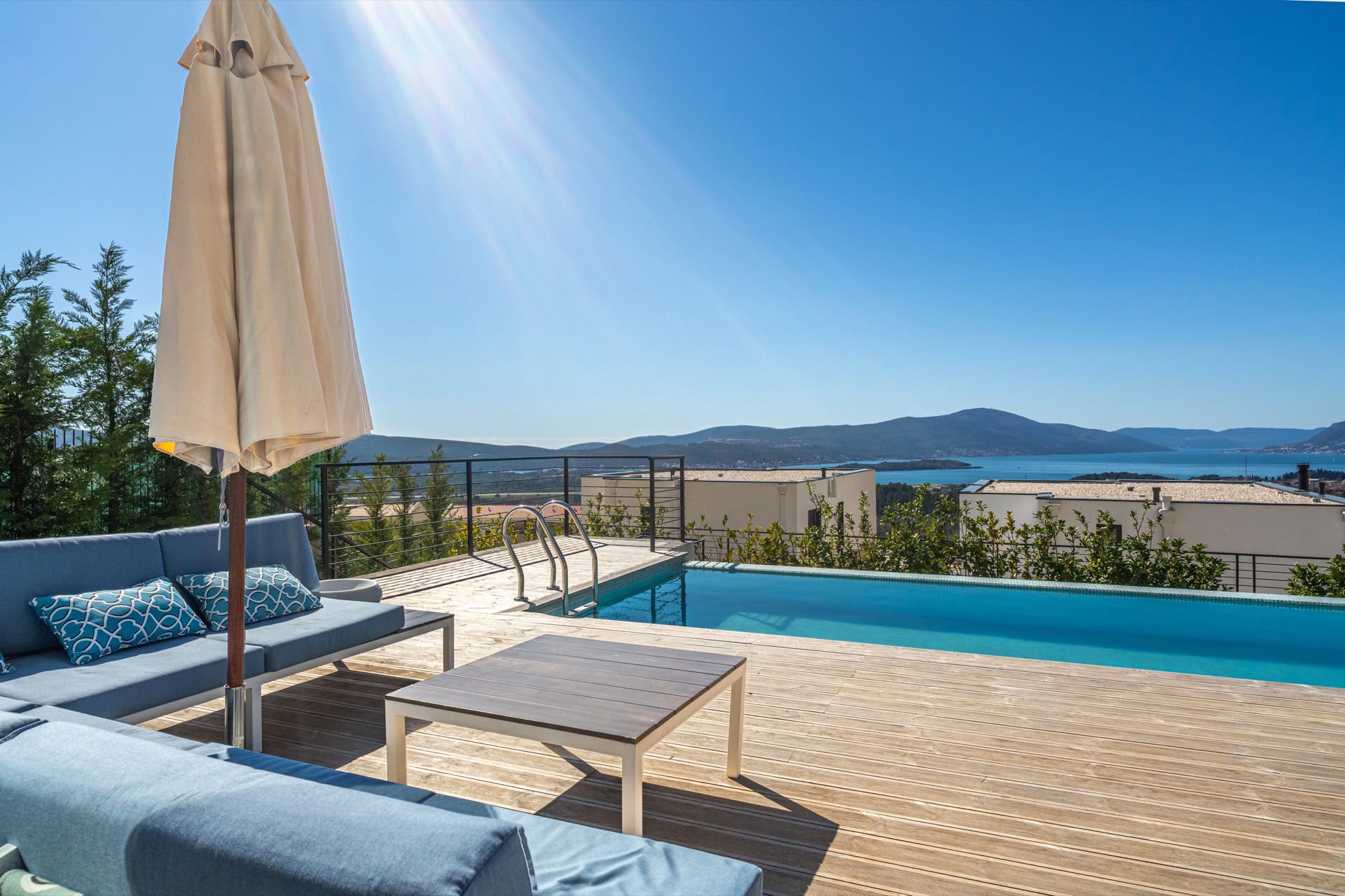 Terrace with a pool and sea view at a villa in Kavac, Montenegro: wooden deck, blue sofas, and a sun umbrella.