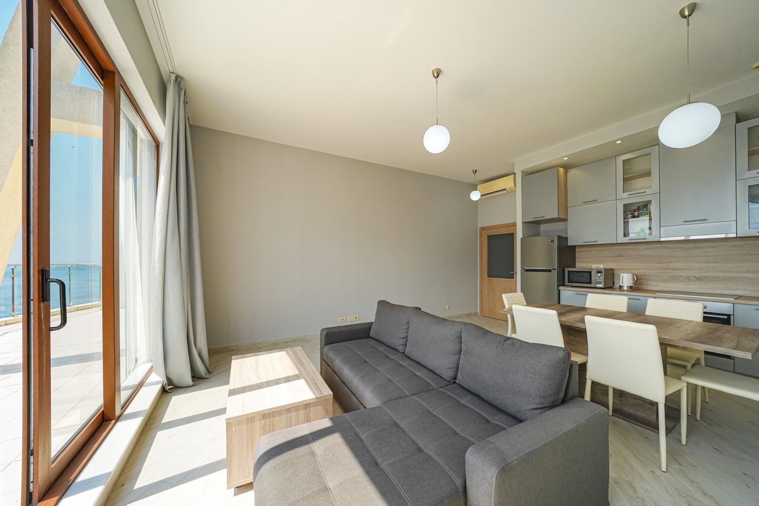 Dnevni boravak sa kuhinjom u stanu u Bečićima, Crna Gora: siva sofa, trpezarija i panoramski pogled na more sa terase.