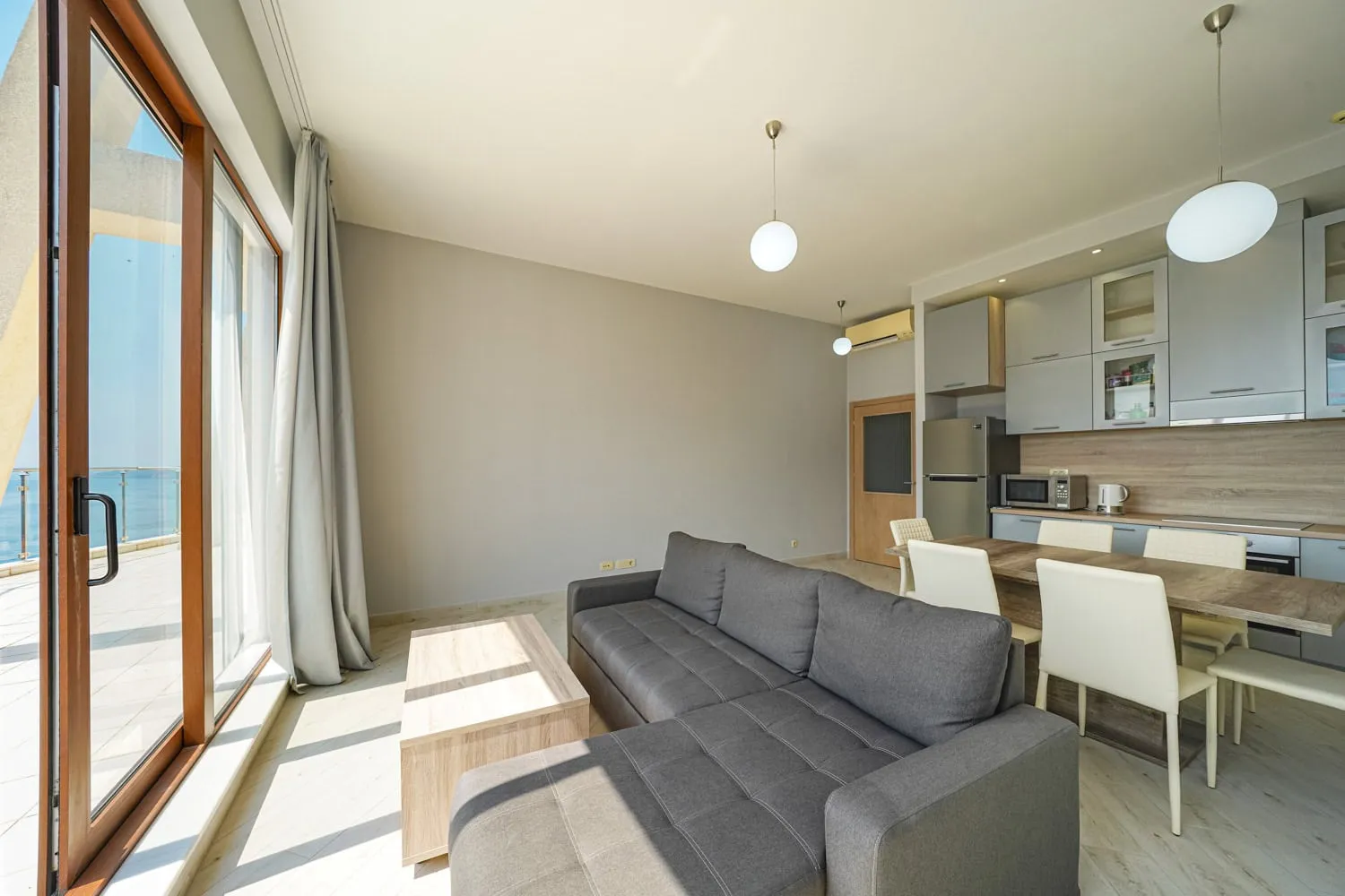 Salon z szarym sofa, drewnianym stołem i otwartą strefą kuchenną w tle przy dużym oknie.