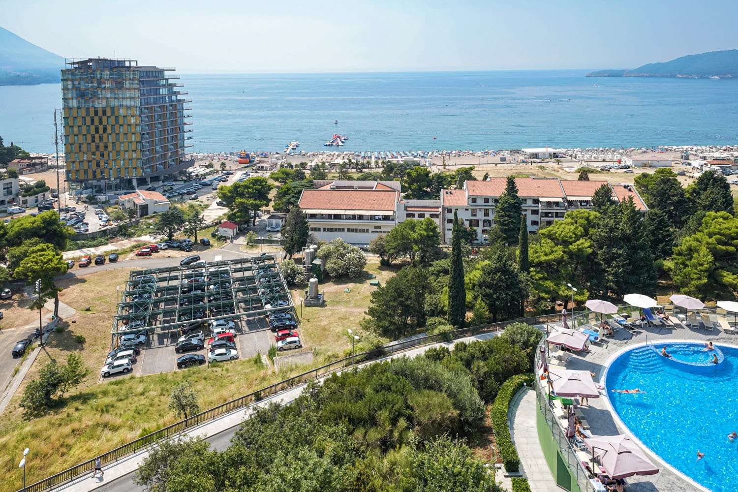 Pogled na more, plažu i bazen sa terase apartmana u Bečićima, Crna Gora. Panorama letovališta sa hotelima i parkingom.