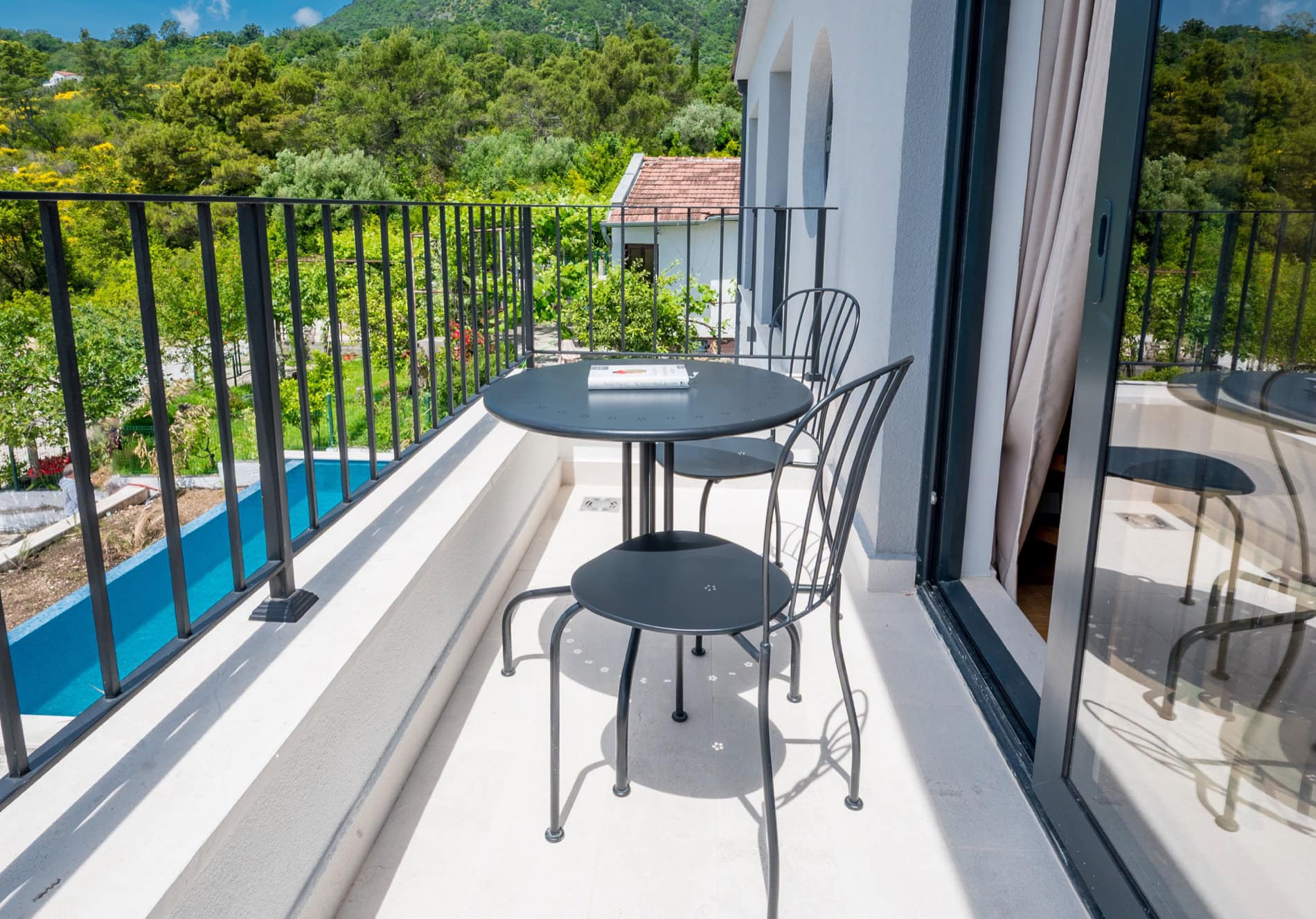 Balkon der Villa in Tivat, Montenegro: schmiedeeiserne Möbel, Blick auf den Pool und den grünen Garten, Panoramaverglasung.