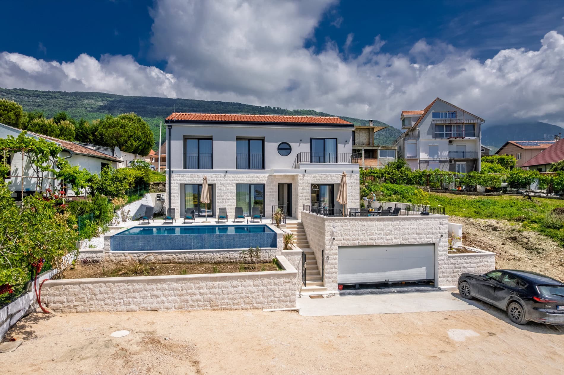 Moderne Villa in Tivat, Montenegro: Steinverkleidung, Pool, Terrasse und Garage vor Bergkulisse.