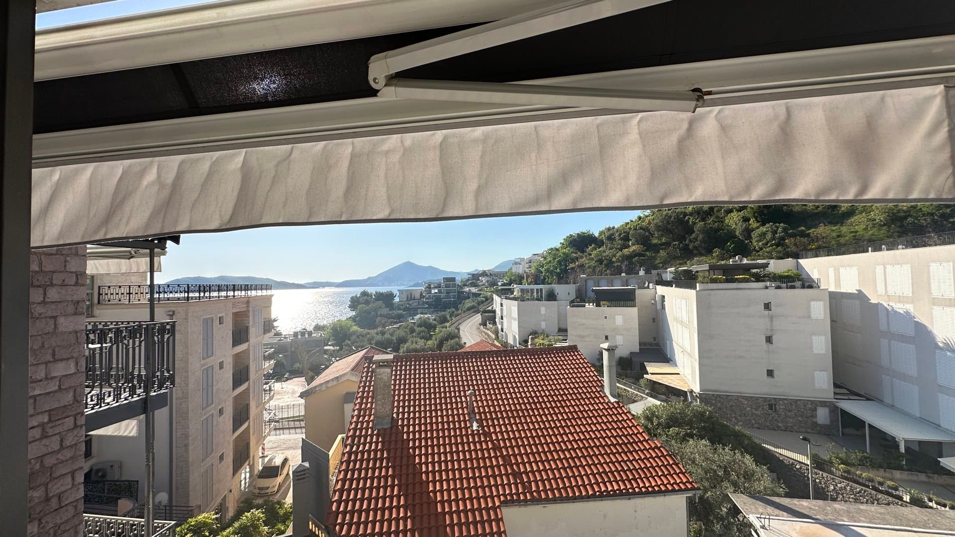 View from the hotel balcony in Pržno, Montenegro: a panorama of the sea, mountains, and red-tiled house roofs on a sunny day.